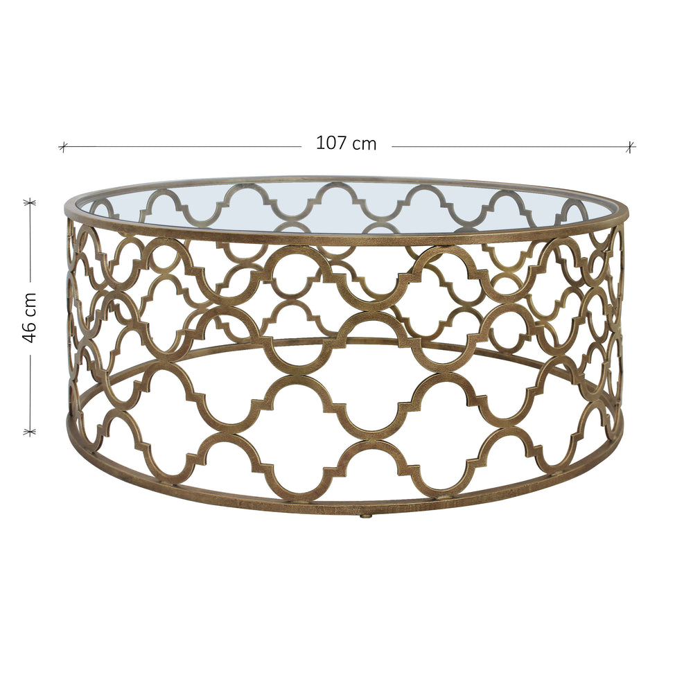 A modern metal coffee table with a quatrefoil geometric pattern in antique gold finish topped with clear glass