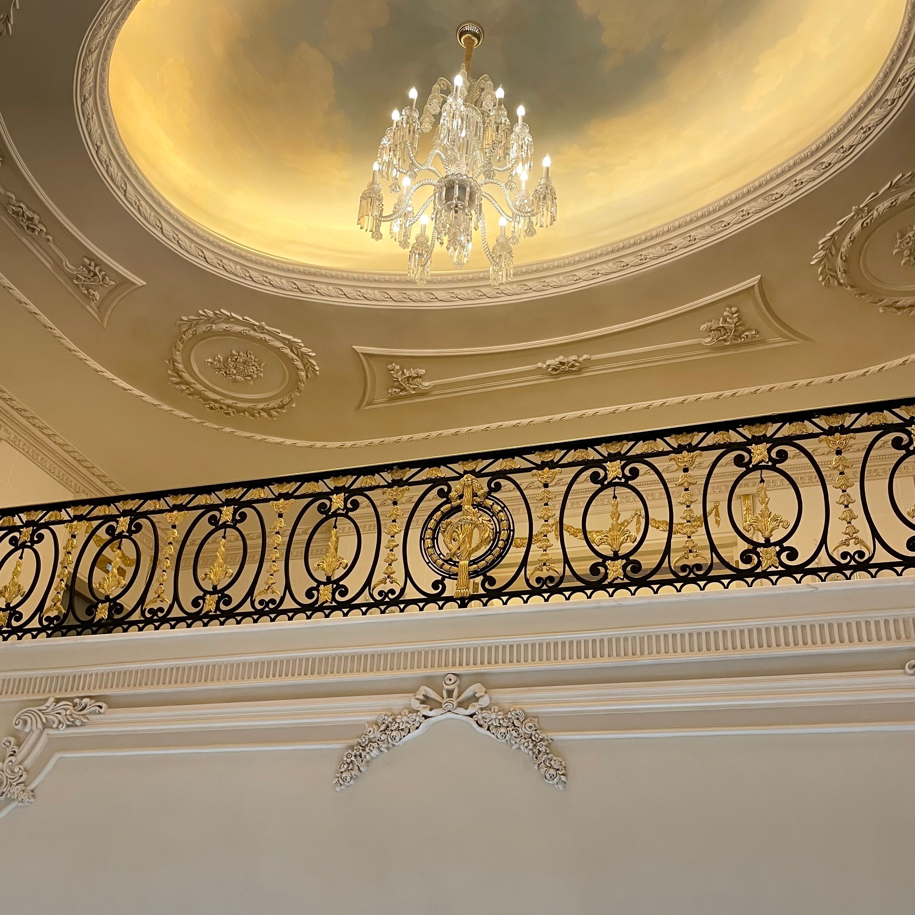 Ornate black and gold wrought iron handrail by Choufani, set against a classical interior with intricate ceiling moldings, a painted dome, and a crystal chandelier.