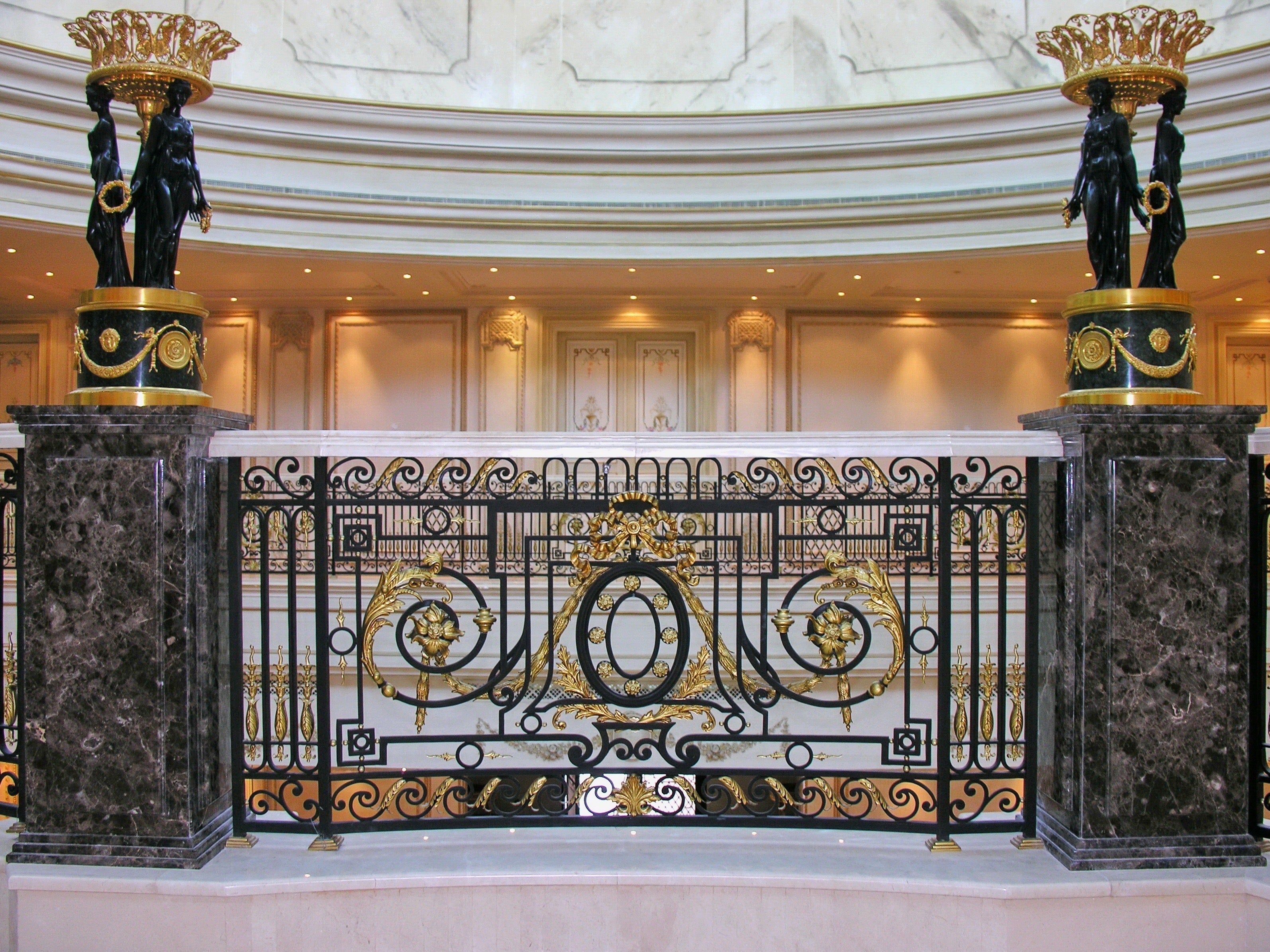 Grand wrought iron handrail with gold detailing and ornamental scrollwork, framed by dark marble columns and classical sculptures in a luxurious interior setting.