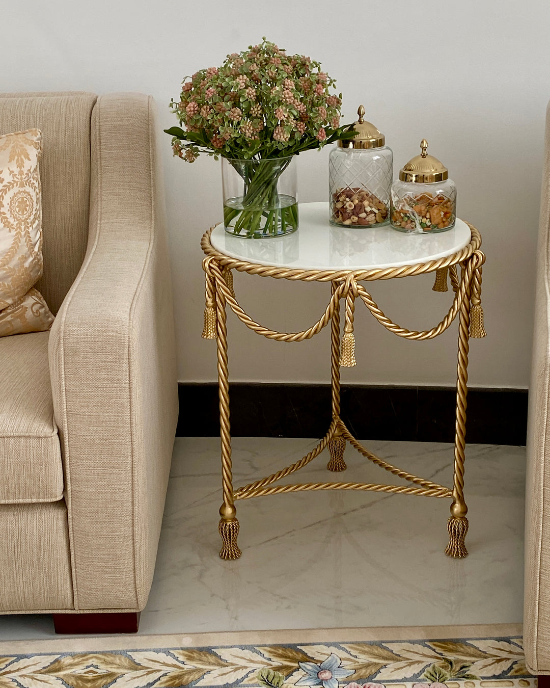 An ornate gold metal side table with a white marble top, styled with a vase of flowers and two glass jars, positioned next to a beige armchair in a living room setting.