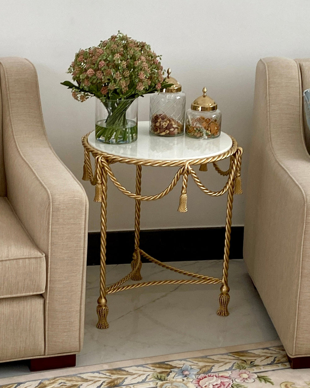 A round side table with a white marble top and ornate gold rope-style legs, styled between two beige armchairs on a patterned rug. The table holds a green glass vase with dried flowers and two glass jars with gold lids.