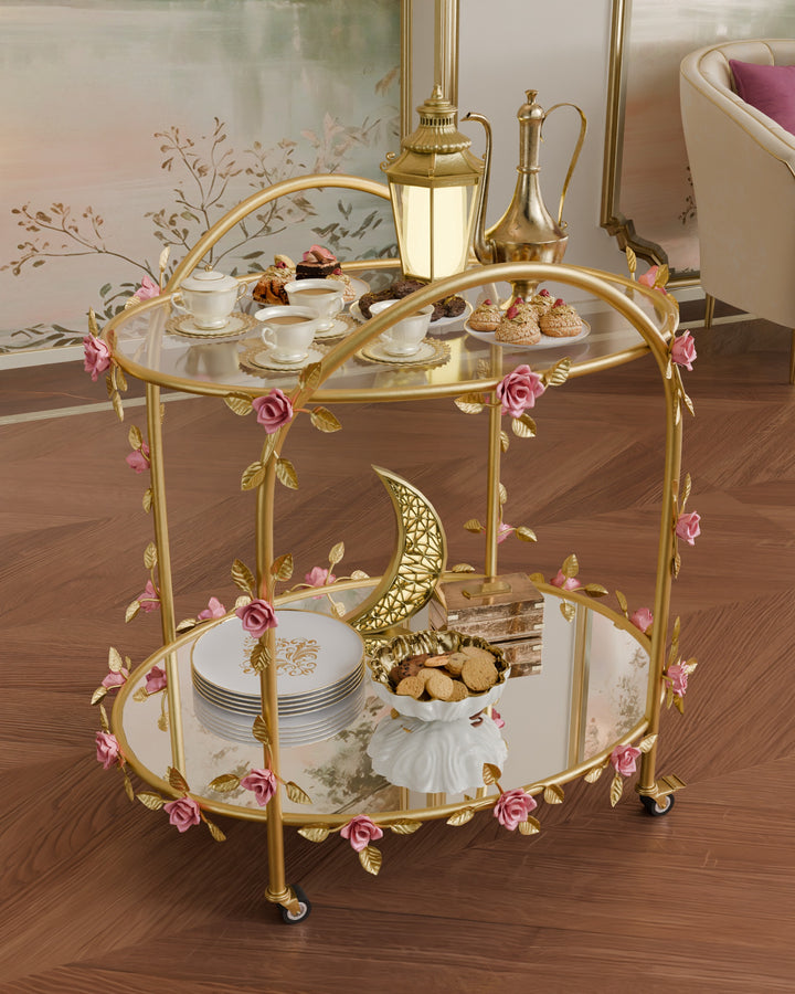 Angled view of Rosetta rose serving trolley in gold wrought iron with pink floral accents and glass shelves, styled with coffee set and desserts in an elegant interior.