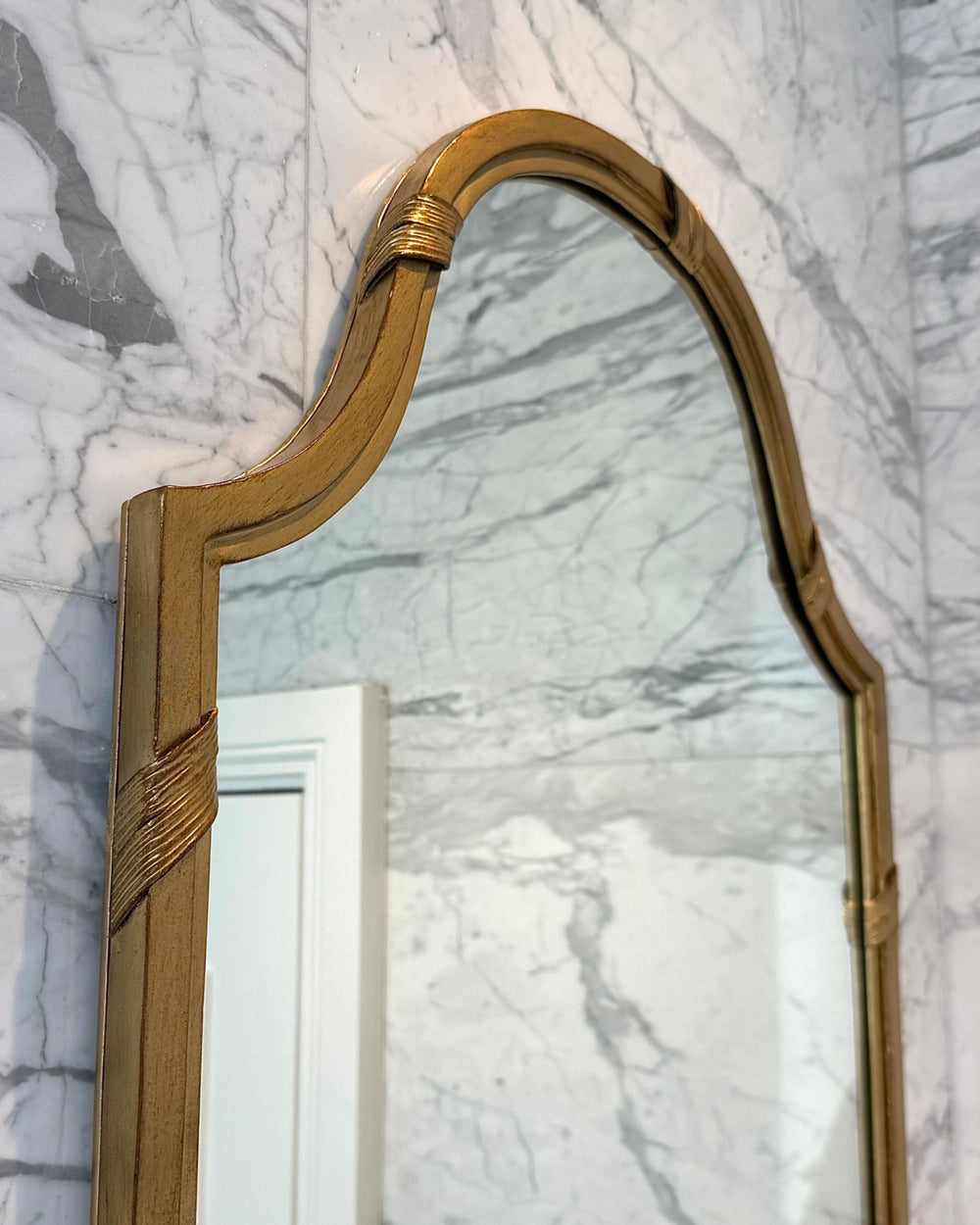 Top detail of the Audrey Mirror’s curved arch and wrapped gold accents, highlighting the craftsmanship against a veined marble wall.