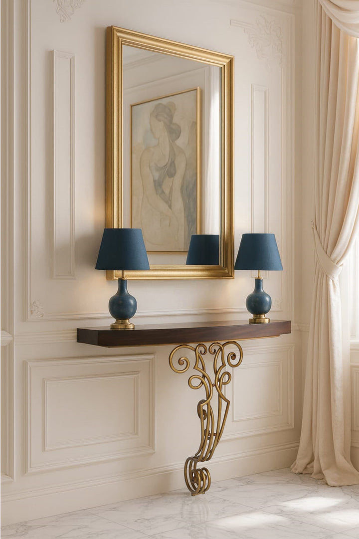 Elegant wall-mounted console with sculpted gold scroll leg, dark wood top, and matching gold-framed mirror styled in a classical cream interior with blue table lamps