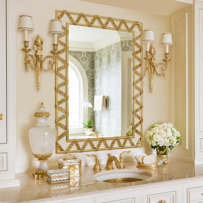 A rectangular golden mirror with a ribbon-like metal strip that forms a zigzag pattern along its perimeter; hangs above a wash basin