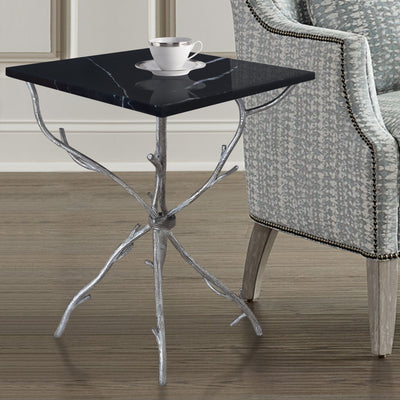 A unique side table with branch-like silver legs topped with a square piece of black marble, stands beside a blue armchair 