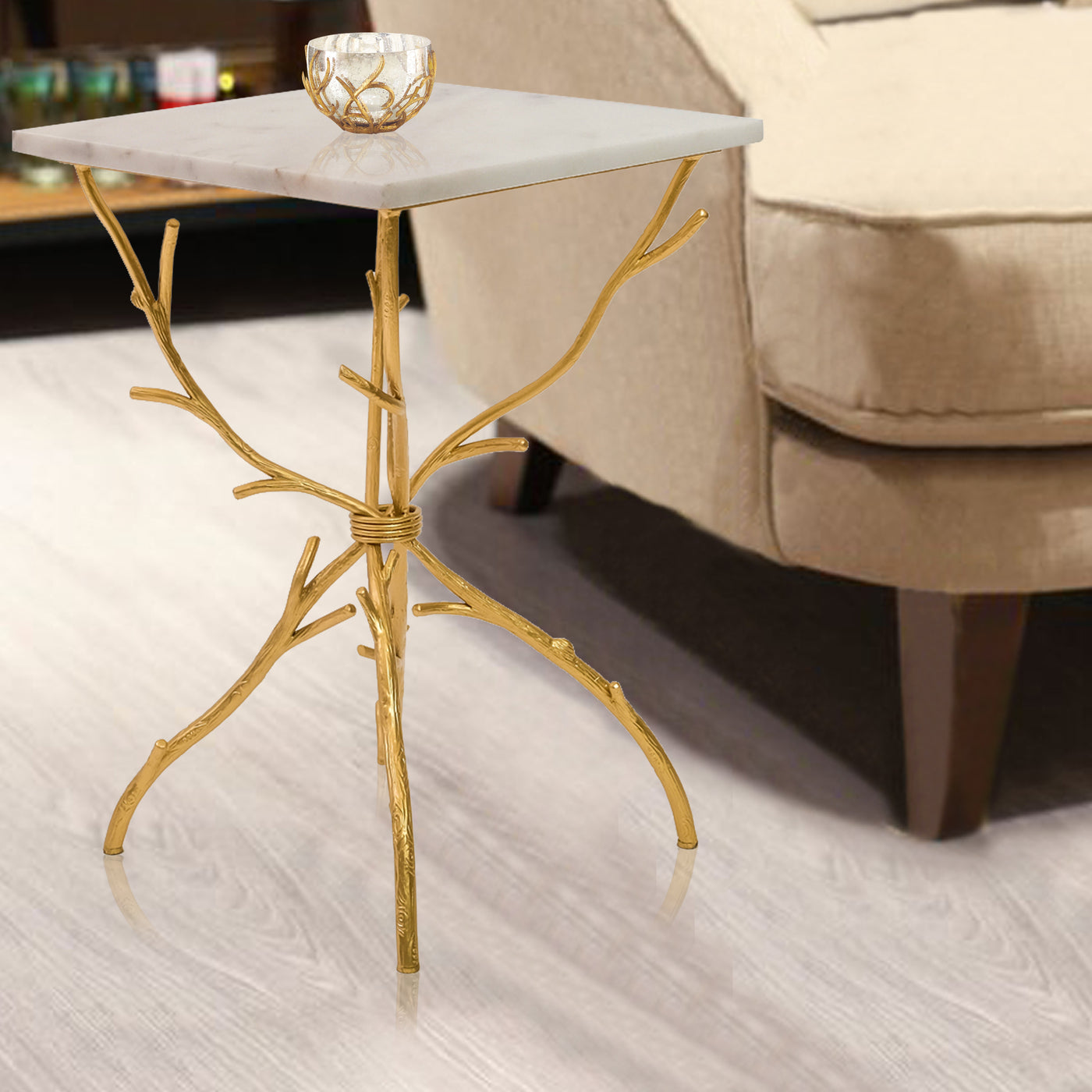 A unique side table with branch-like golden legs carry a square white piece of marble, stands beside an armchair 