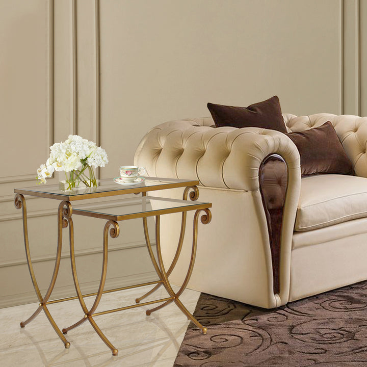 A set of two wrought iron nesting tables with long scrolled legs painted in antique gold and topped with glass sit beside a contemporary armchair