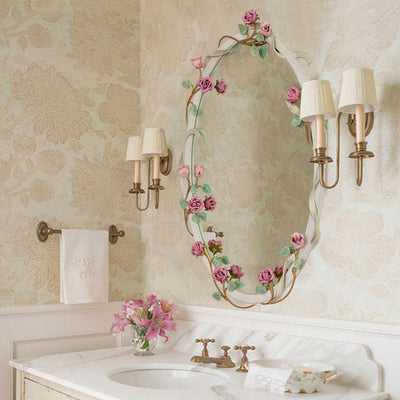 An oval shaped mirror with a wreath of pink roses and green leaves wrapped along its border, hangs above a bathroom sink